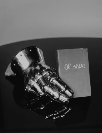 Close-up studio shot of a metal gauntlet resting next to a book titled 'Orlando' on a reflective black surface, creating a dramatic still life.