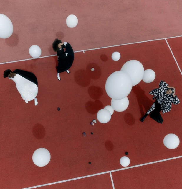 Three adults in black and white outfits are positioned among white balloons on a coral-colored outdoor court, captured in an eye-level shot.