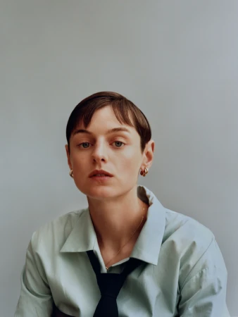 Portrait of a woman with short dark hair wearing a light blue collared shirt and black tie against a grey background.