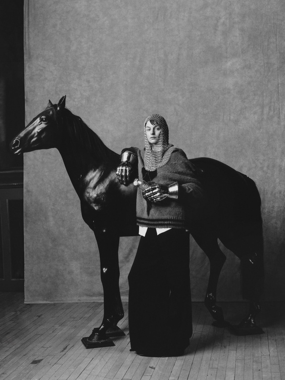 Monochrome portrait of a person in chainmail, posing with a black horse in an indoor setting with wood floors and a neutral backdrop.
