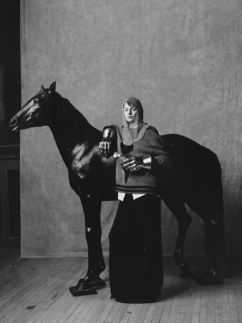 Monochrome portrait of a person in chainmail, posing with a black horse in an indoor setting with wood floors and a neutral backdrop.