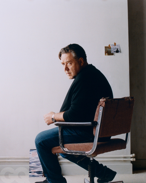 Middle-aged man sits in a vintage office chair indoors with a blank background, wearing a black shirt and dark jeans.