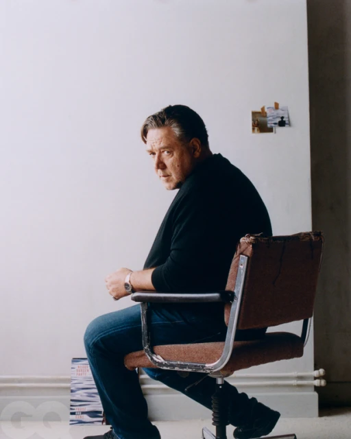 Middle-aged man sits in a vintage office chair indoors with a blank background, wearing a black shirt and dark jeans.