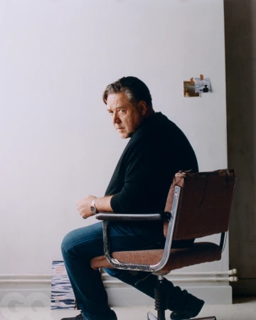 Middle-aged man sits in a vintage office chair indoors with a blank background, wearing a black shirt and dark jeans.