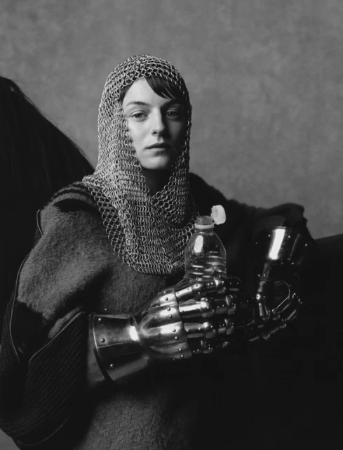 A woman in chainmail and metal gloves holds a water bottle in a dramatic black and white photograph, seated against a grey backdrop.