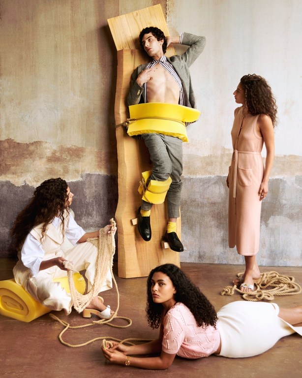 Fashion shoot with a bare-chested man strapped to wood and three women wearing dresses and knitwear in a studio setting.