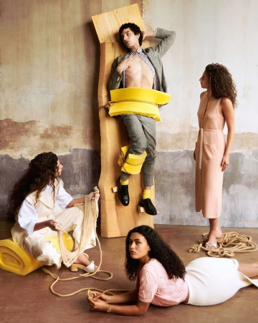 Fashion shoot with a bare-chested man strapped to wood and three women wearing dresses and knitwear in a studio setting.