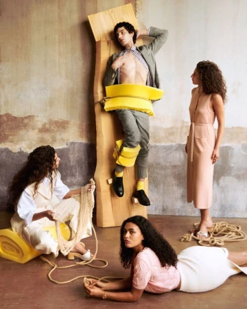 Fashion shoot with a bare-chested man strapped to wood and three women wearing dresses and knitwear in a studio setting.