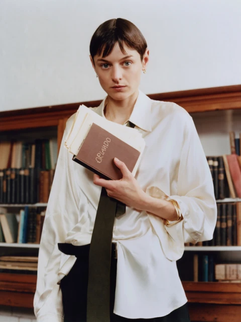 Indoor portrait of a brunette adult female in a satin shirt, holding books in a library setting with bookshelves. Editorial fashion.
