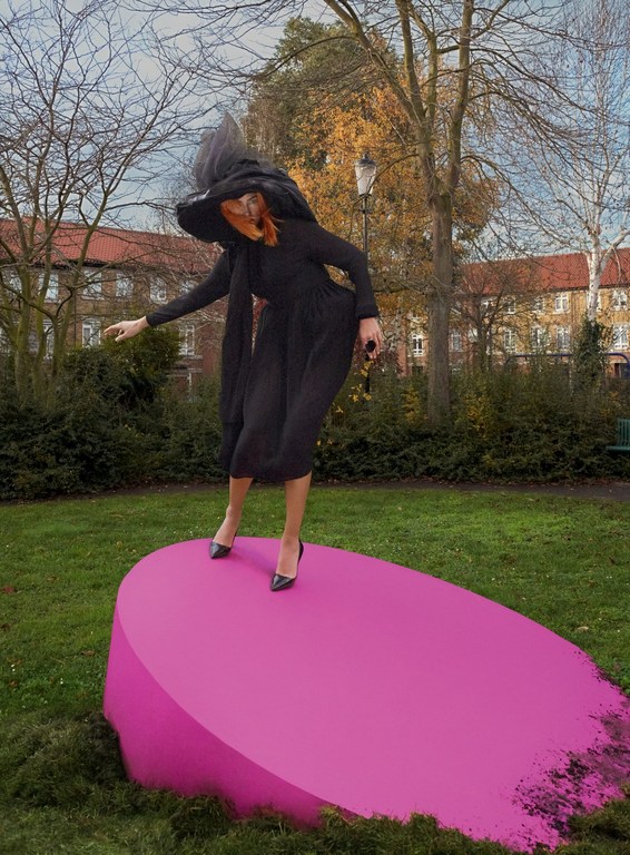 A woman in a witch costume is standing on a large, bright pink oval platform in a park setting, with trees and buildings in the background.