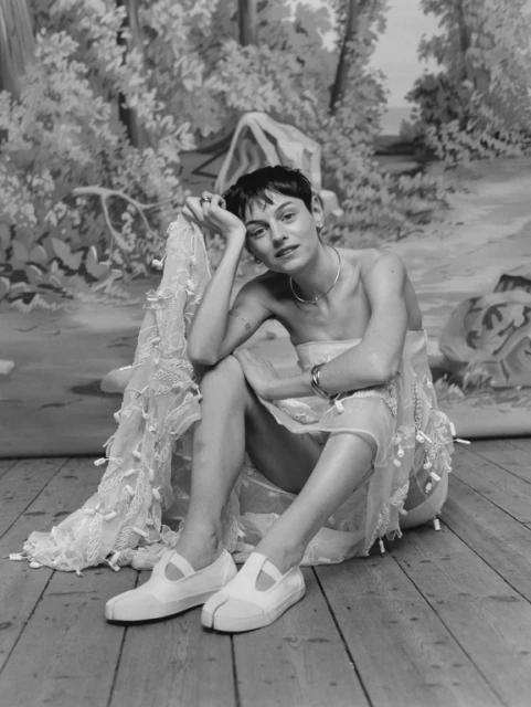 Monochrome portrait of a young woman with a pixie cut sitting in a light-colored dress and white shoes, with a painted backdrop.