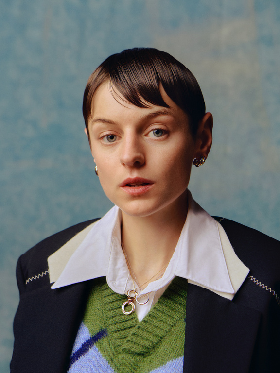 Portrait of a person with short dark hair wearing a white collared shirt, green patterned sweater vest, and dark blazer.