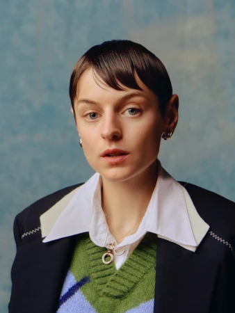 Portrait of a person with short dark hair wearing a white collared shirt, green patterned sweater vest, and dark blazer.