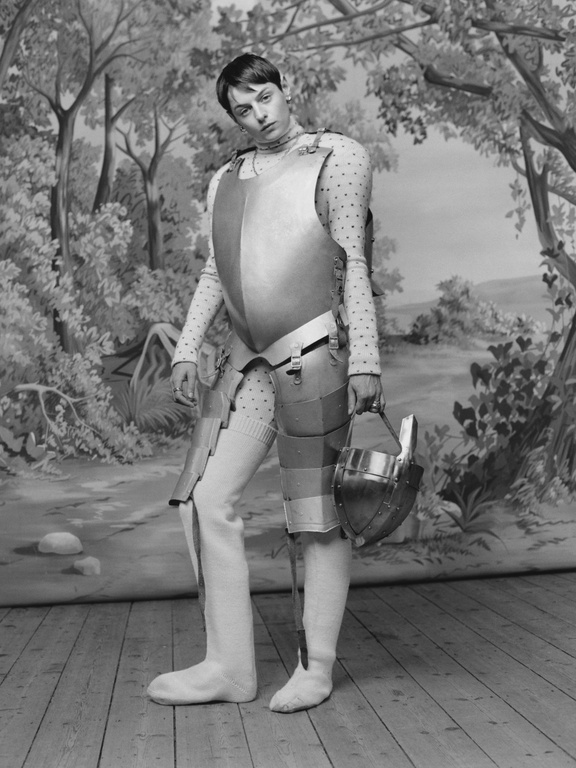 Person in full silver armour with helmet stands against painterly backdrop featuring trees and landscape. Full shot in desaturated tones.