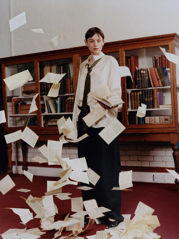 Indoor fashion shot of a female adult standing amidst scattered papers in front of a bookcase, wearing a white shirt and dark skirt.