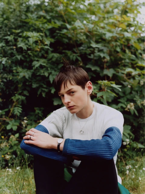 Young adult with short hair poses outdoors in nature, wearing a striped sweater and dark pants.  Wristwatch and rings visible.