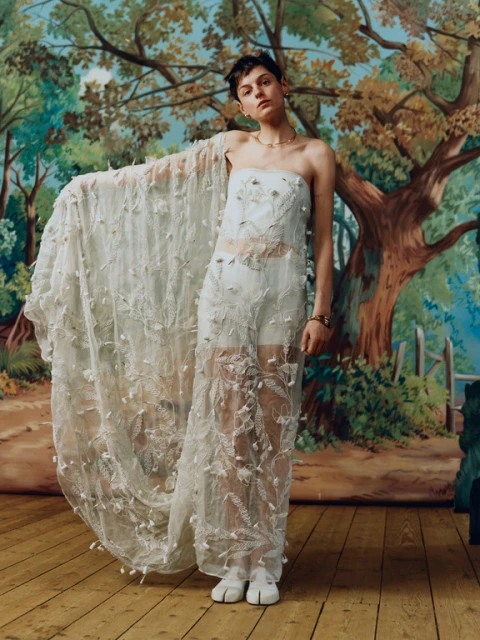 A person wearing a sheer dress with floral details poses in front of a backdrop depicting a forest and trees in an artistic style.