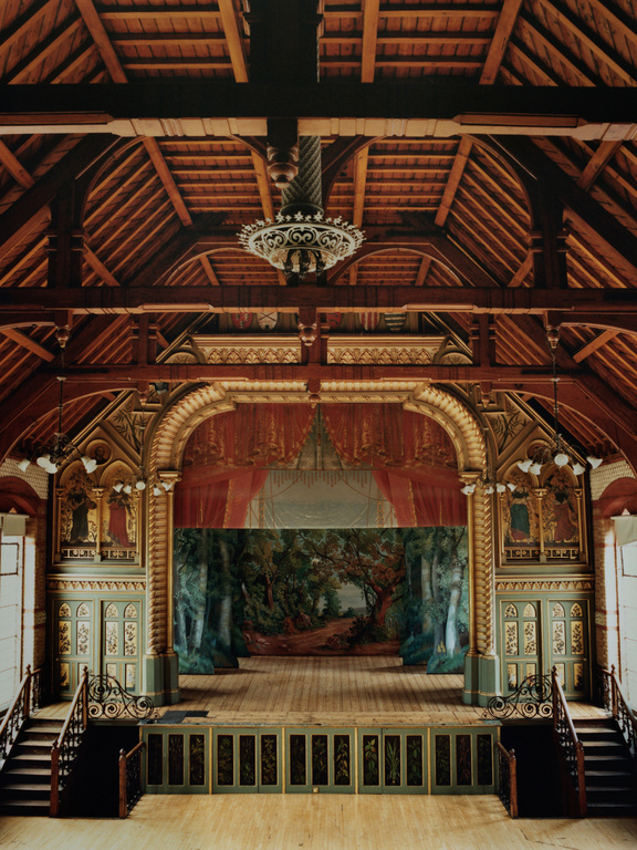 Ornate vintage theater interior with a forest backdrop, wooden stage, decorative arches, a hanging chandelier, and flanking staircases.