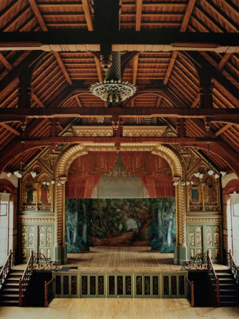 Ornate vintage theater interior with a forest backdrop, wooden stage, decorative arches, a hanging chandelier, and flanking staircases.