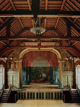 Ornate vintage theater interior with a forest backdrop, wooden stage, decorative arches, a hanging chandelier, and flanking staircases.