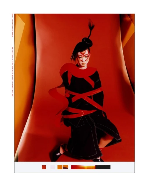 Asian woman in dark dress with red accents poses against vibrant red backdrop, styled with dramatic makeup and a unique updo.