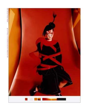 Asian woman in dark dress with red accents poses against vibrant red backdrop, styled with dramatic makeup and a unique updo.