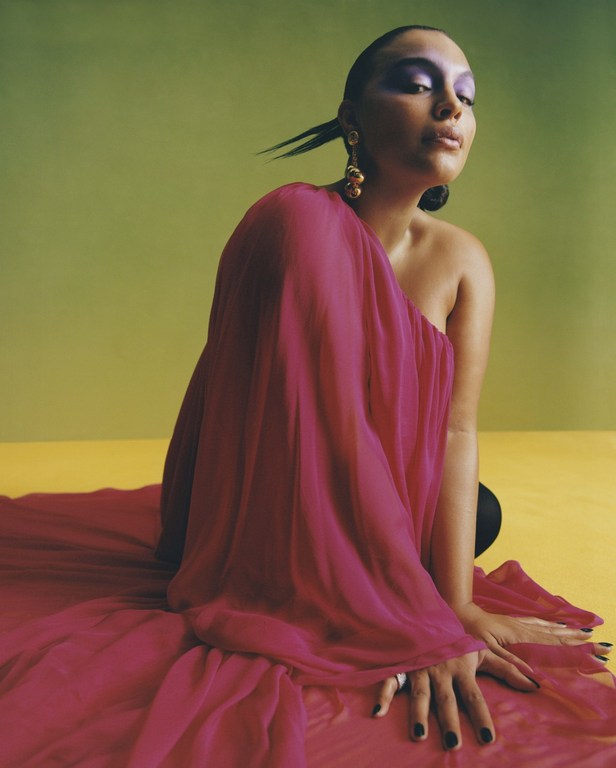 Elegant woman in a fuchsia dress and dramatic makeup kneeling against a bright yellow and green background, highlighted by studio lighting.