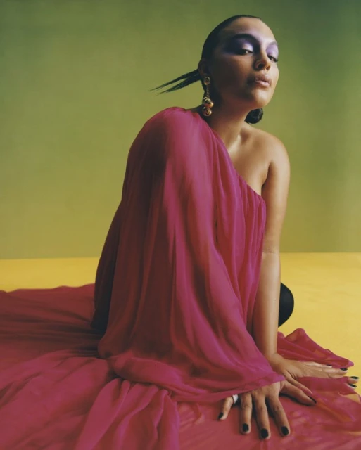 Elegant woman in a fuchsia dress and dramatic makeup kneeling against a bright yellow and green background, highlighted by studio lighting.