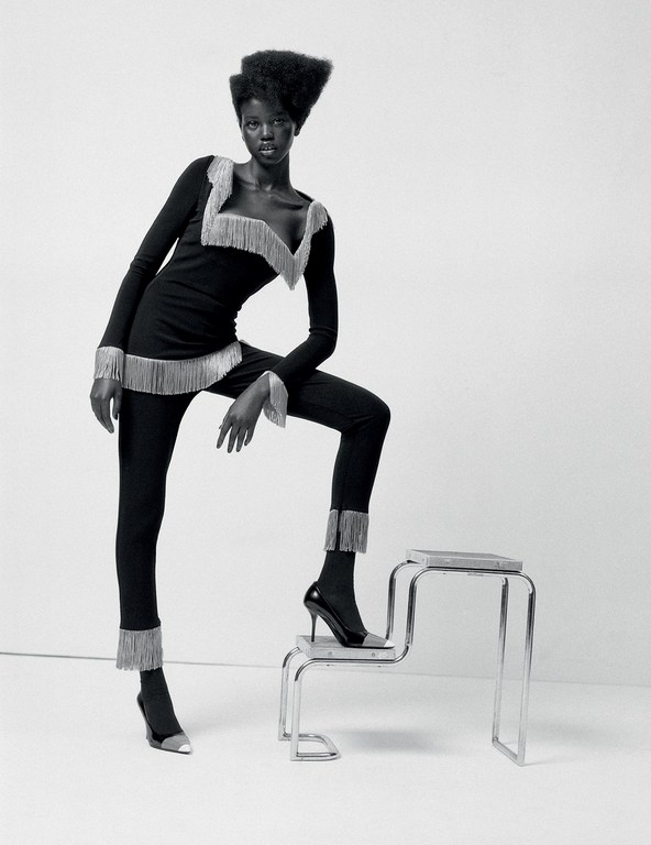 Black and white fashion shot of an African woman in a long-sleeved black outfit with silver fringe, posing on a silver metal step stool.