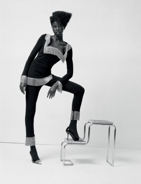 Black and white fashion shot of an African woman in a long-sleeved black outfit with silver fringe, posing on a silver metal step stool.