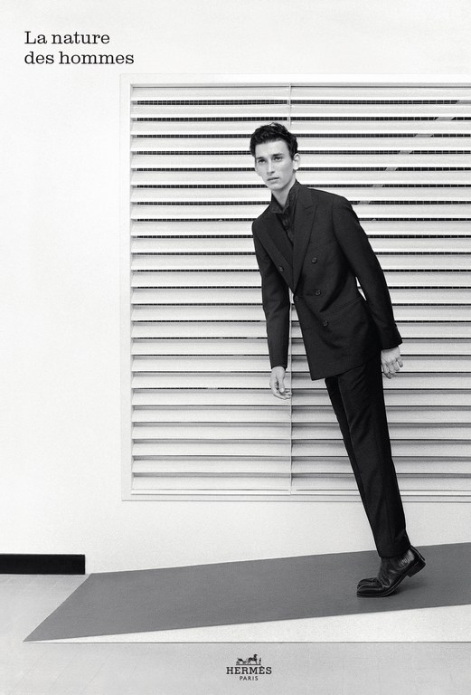 Monochromatic studio shot of a male model wearing a dark suit, posing against a white backdrop with horizontal window blinds and gray flooring.