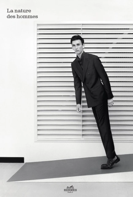 Monochromatic studio shot of a male model wearing a dark suit, posing against a white backdrop with horizontal window blinds and gray flooring.