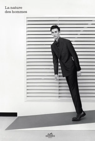 Monochromatic studio shot of a male model wearing a dark suit, posing against a white backdrop with horizontal window blinds and gray flooring.