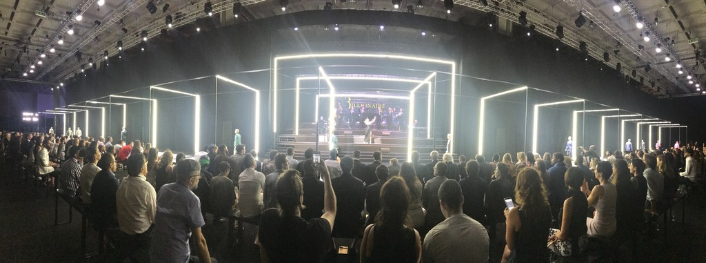 Runway show at fashion week featuring models and audience in large event space with bright lighting, dark walls, and a raised stage.