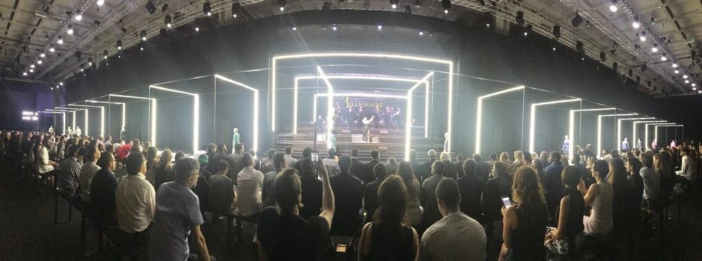 Runway show at fashion week featuring models and audience in large event space with bright lighting, dark walls, and a raised stage.