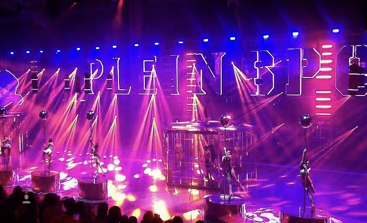 A performance stage featuring dancers on poles with dramatic lighting and large text backdrop, creating a vibrant and dynamic visual.