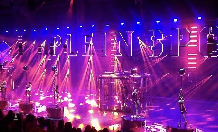 A performance stage featuring dancers on poles with dramatic lighting and large text backdrop, creating a vibrant and dynamic visual.