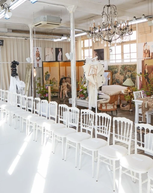 Fashion show setup with rows of white chairs on a white floor, chandelier, artwork, and camera in an industrial-style space.