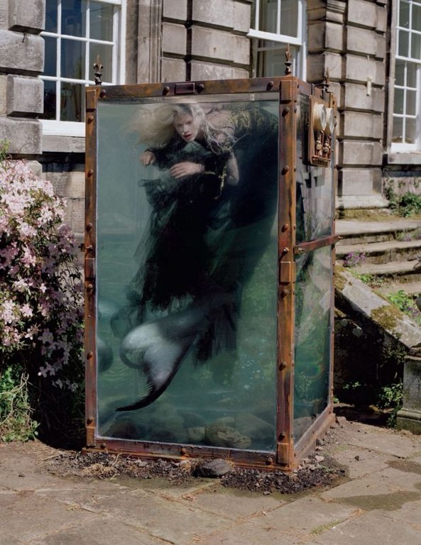 Artistic scene of a blonde woman in a black dress inside a glass tank filled with water, set outdoors near a stone building and plants.