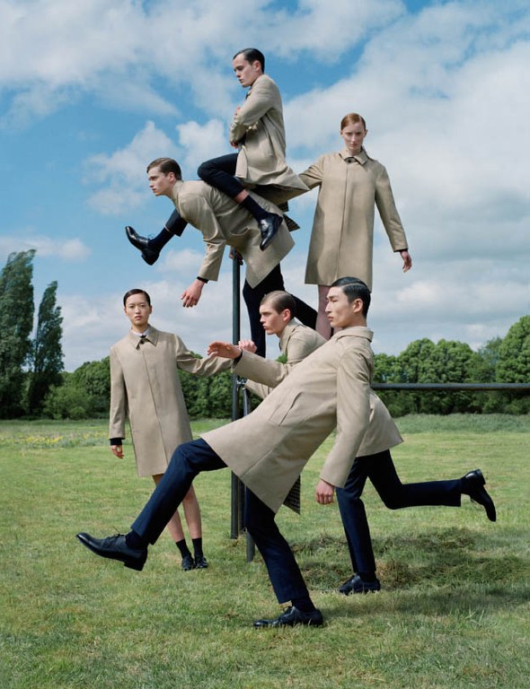 Outdoor fashion shoot with six adult models in tan trench coats and dark pants, arranged in a whimsical human sculpture against a blue sky.