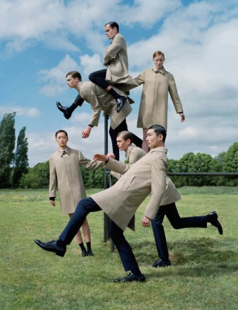 Outdoor fashion shoot with six adult models in tan trench coats and dark pants, arranged in a whimsical human sculpture against a blue sky.
