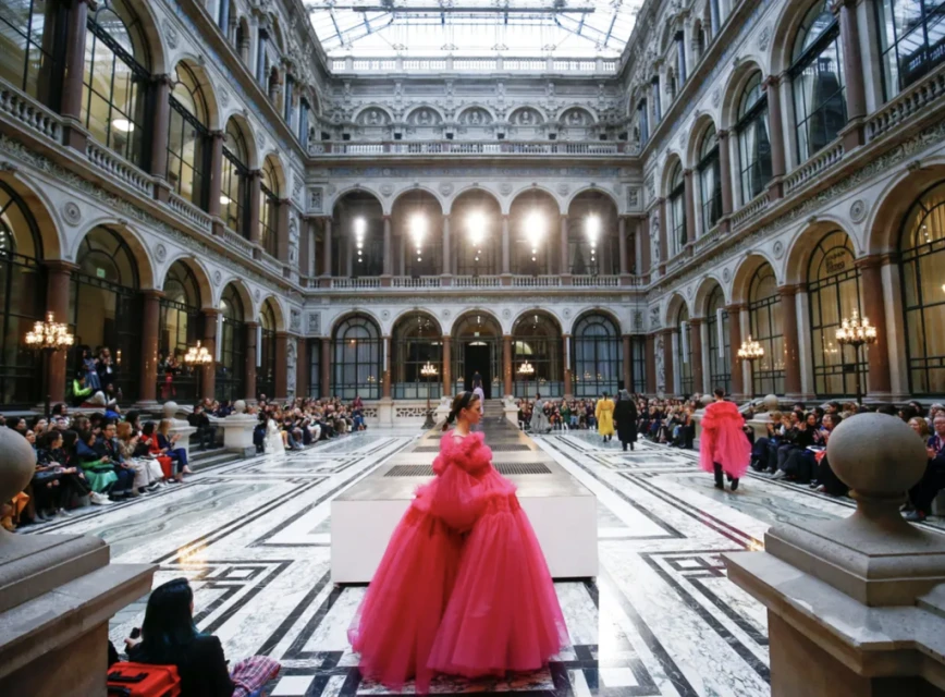 Runway fashion show in an opulent hall: A model in a vibrant pink gown walks a marble runway as spectators watch from the sides.