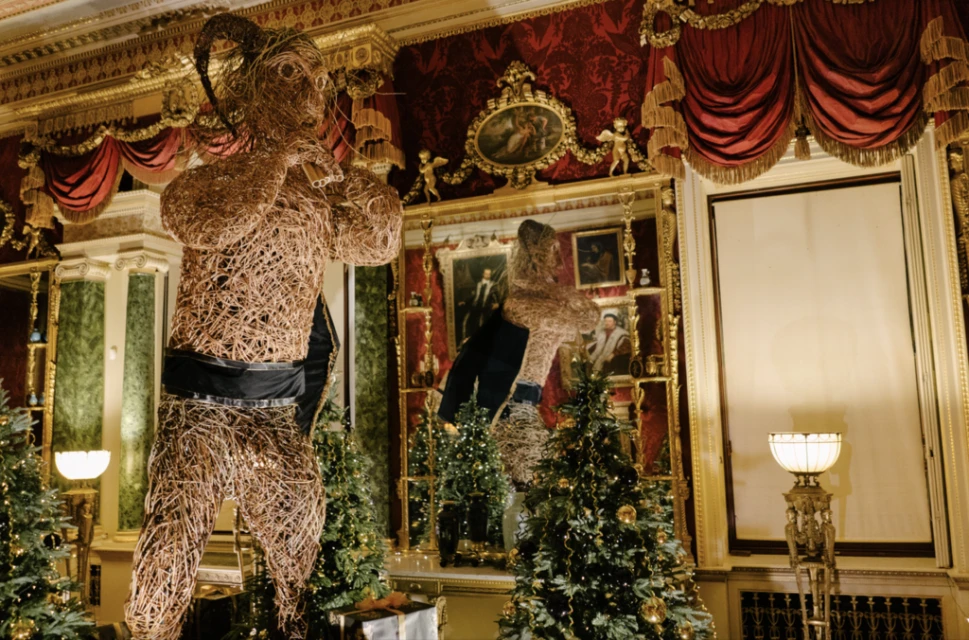 Intricate woven sculpture of a human figure stands amidst Christmas trees in a grand hall, showcasing artistic design and holiday decor.
