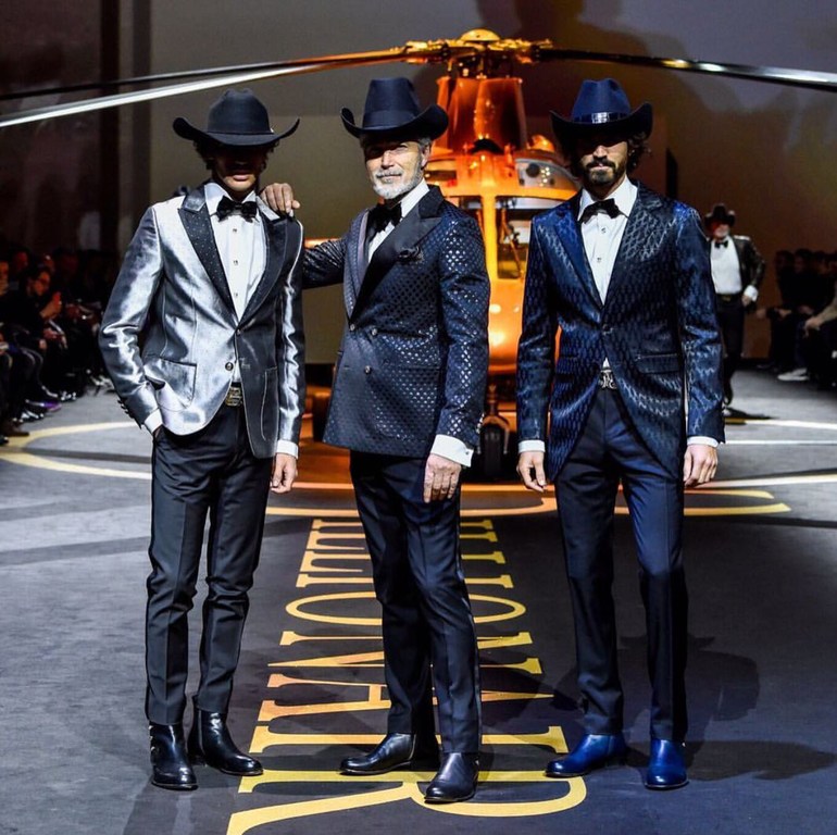 Three adult men in cowboy hats and suits pose on a runway in front of a helicopter, captured in a medium shot in an indoor setting.