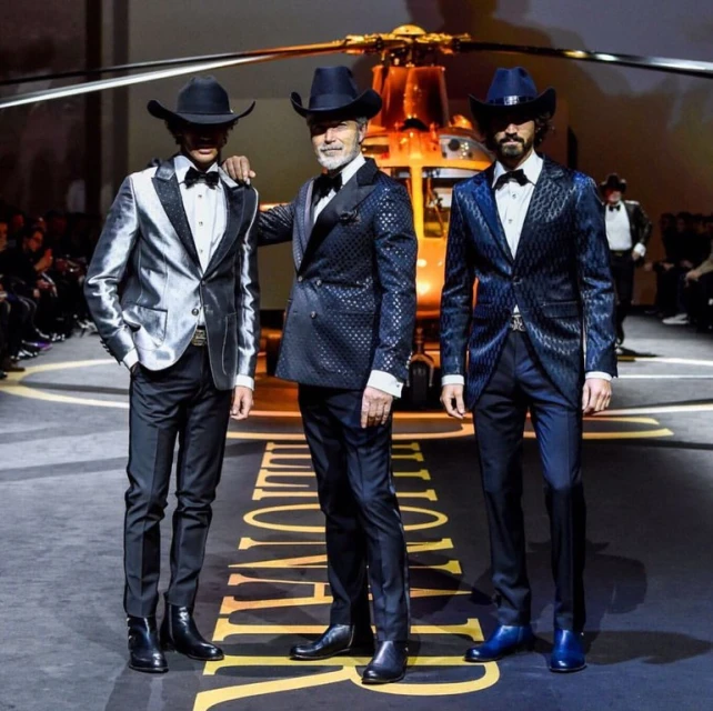 Three adult men in cowboy hats and suits pose on a runway in front of a helicopter, captured in a medium shot in an indoor setting.