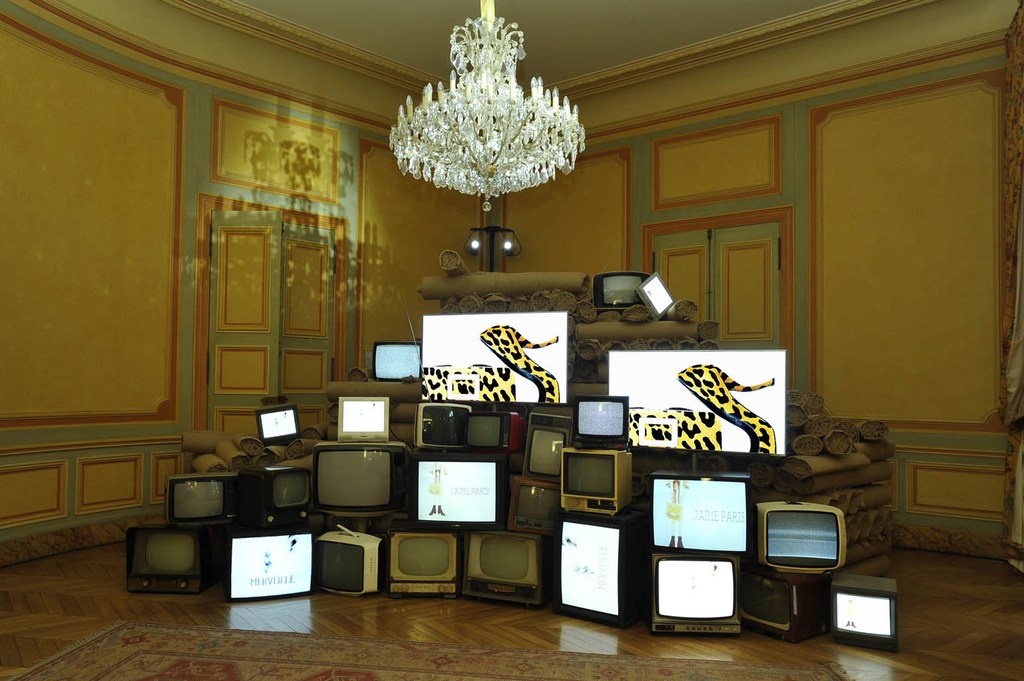 Indoor art installation with stacked retro televisions showing art, in a room with ornate walls, chandelier, and carpet.