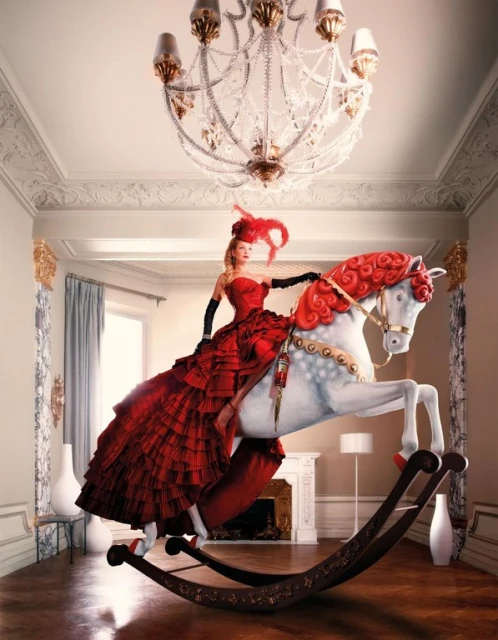 Woman in red ballgown on rocking horse in a room with a chandelier, moldings, and wood floors in an indoor editorial setting.