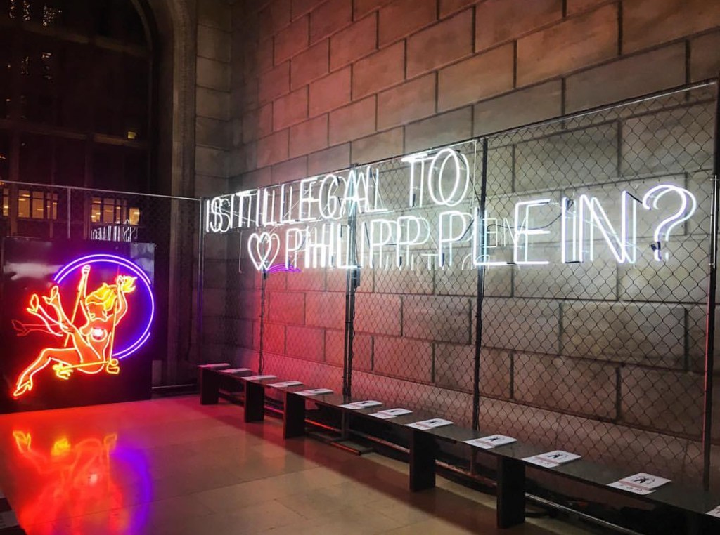 Neon art installation featuring a glowing cartoon figure and text 'IS IT ILLEGAL TO HEART PHILIPP PLEIN?' inside a brick-walled building with benches.