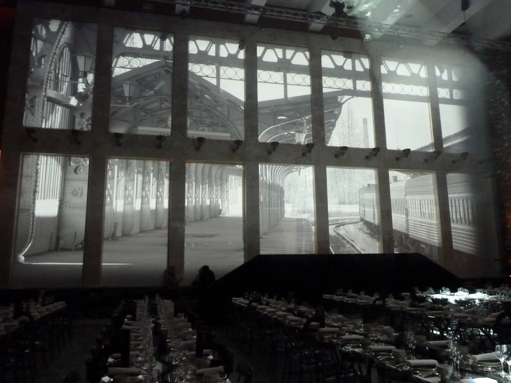 Indoor event with dinner tables and a large backdrop projection of a vintage train station and train in monochrome tones.