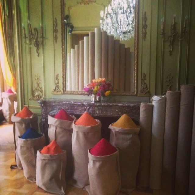 Bags of colorful pigment or dye line a room with decorative molding and a chandelier, rolls of carpet stand against the back wall.